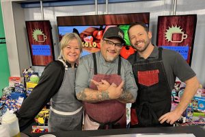 A group of three individuals: two men and one woman. In front of them is a table set up with a pot and many ingredients as they are preparing for a cooking demonstration.